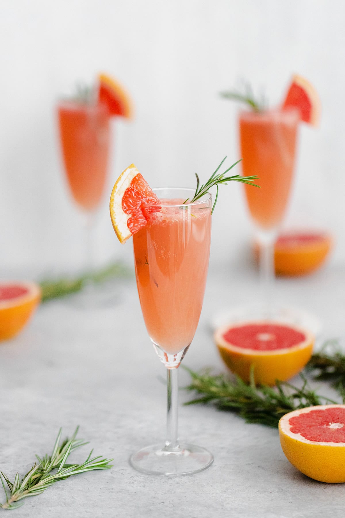 Grapefruit Mimosa in 3 flute glasses garnished with fresh rosemary and a quarter of a slice of grapefruit on the rim. Grapefruits as decoration in the background.