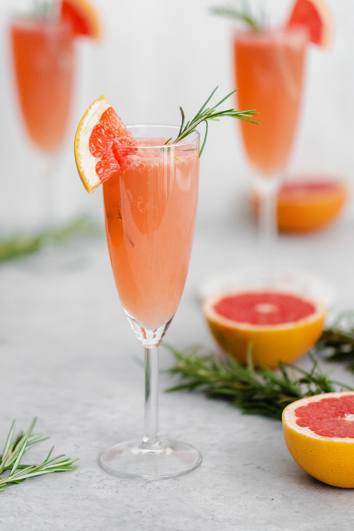 Grapefruit Mimosa in 3 flute glasses garnished with fresh rosemary and a quarter of a slice of grapefruit on the rim. Grapefruits as decoration in the background.