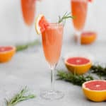 Grapefruit Mimosa in 3 flute glasses garnished with fresh rosemary and a quarter of a slice of grapefruit on the rim. Grapefruits as decoration in the background.