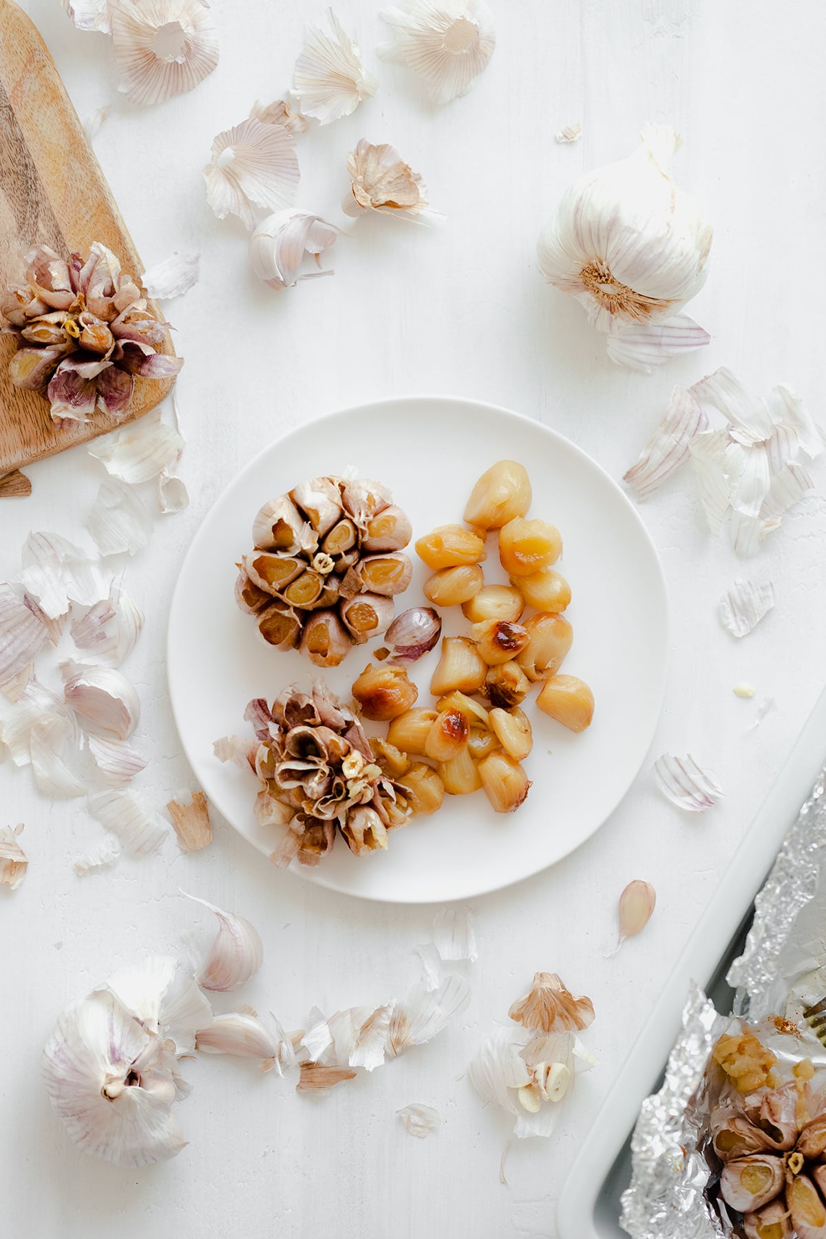 A white plat on white background with roasted garlic on it. Two bulbs, one full, one empty with the roasted garlic cloves on the right side of the plate.