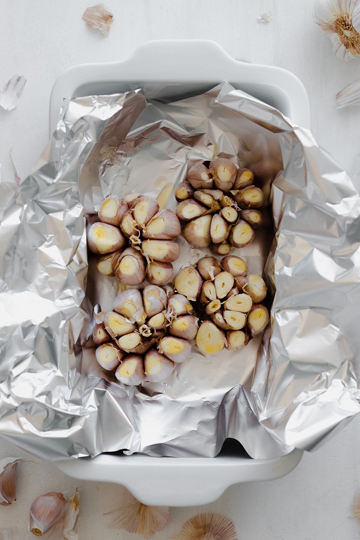 Four whole garlic bulbs on foil in a white baking dish. White background, overhead shot.