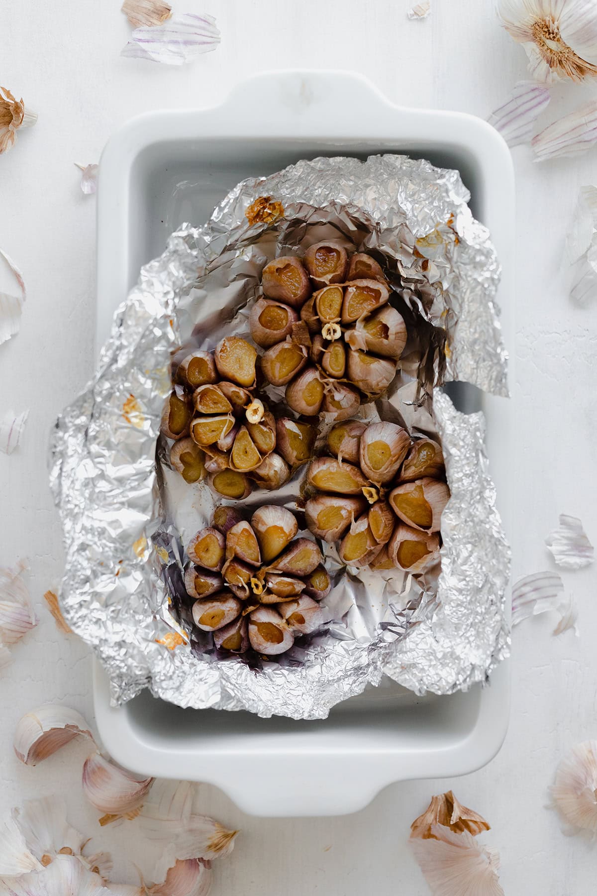 Roasted garlic bulbs unwrapped in foil in a white baking dish on a white background.