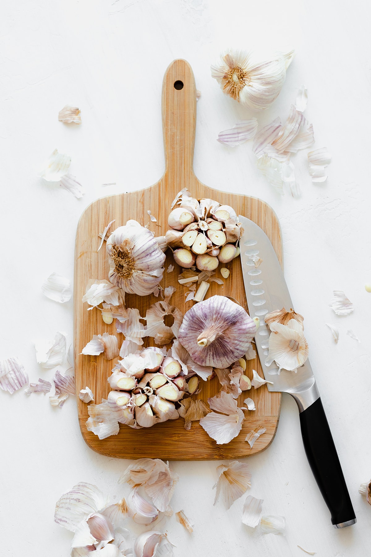 A photo of 4 garlic bulbs, two with the tops cut off, on a wooden cutting board with the knife of the right side of it. On a white background.