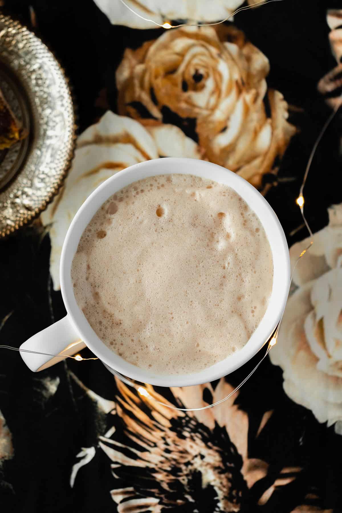 Blossoming Rose Tea Latte in a white mug. No garnish. On a black table cloth with beige rose print. Fairy lights around mug.