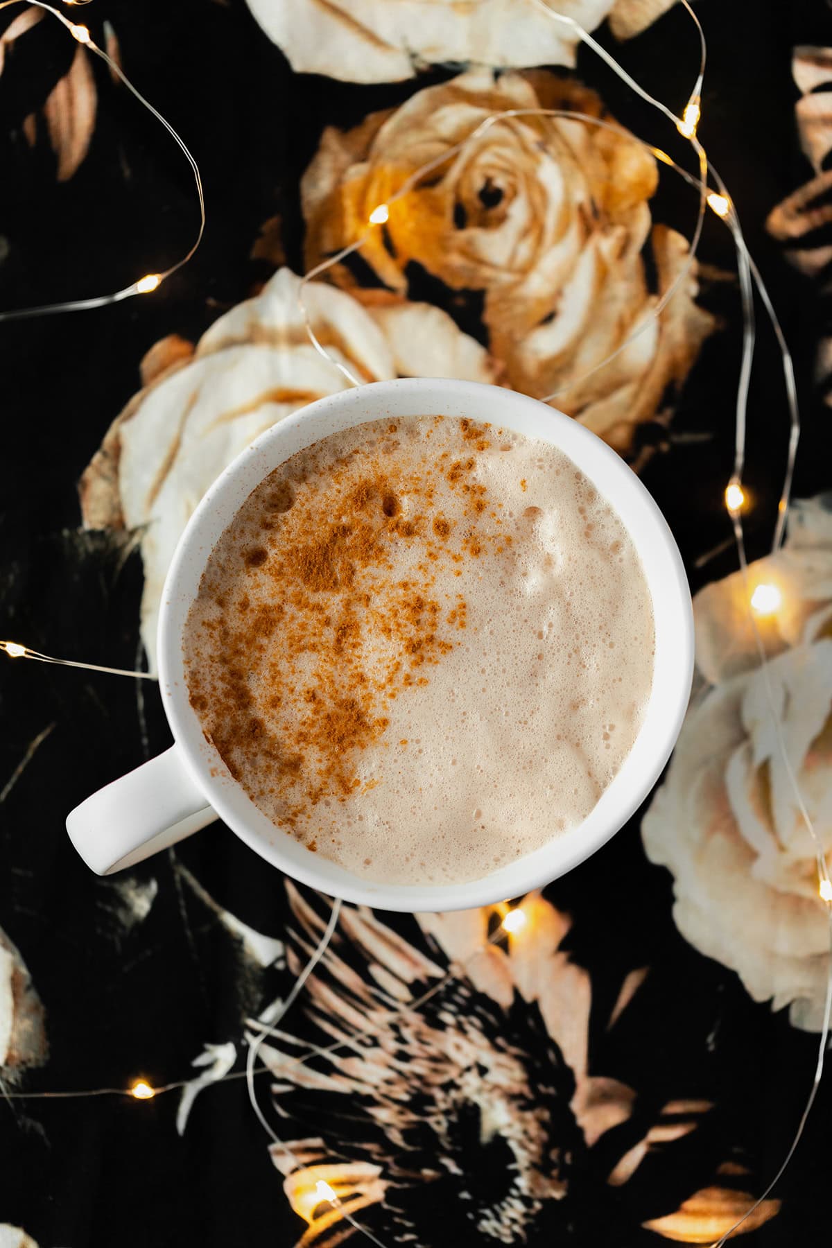 Blossoming Rose Tea Latte in a white mug with a light dusting of cinnamon on the left side. On a black table cloth with beige rose print. Fairy lights around mug.