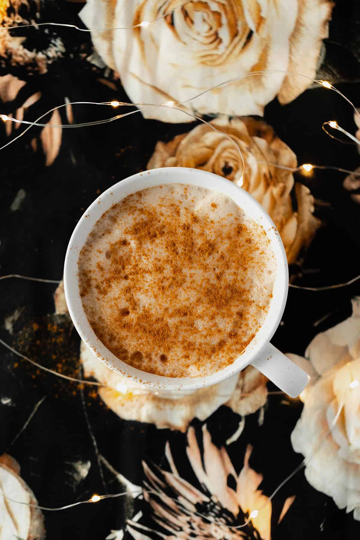 Blossoming Rose Tea Latte in a white mug with a heavy dusting of cinnamon all over. On a black table cloth with beige rose print. Fairy lights around mug.