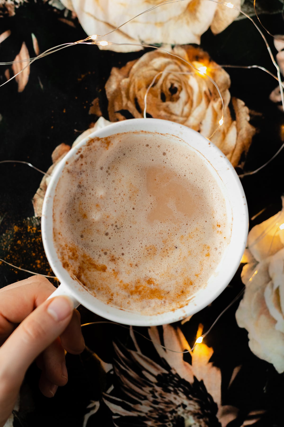 A hand holding a half drunk latte in a white mug with a light dusting of cinnamon on the left side. On a black table cloth with beige rose print. Fairy lights around mug.
