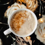 Blossoming Rose Tea Latte in a white mug with a light dusting of cinnamon on the left side. On a black table cloth with beige rose print. Fairy lights around mug.