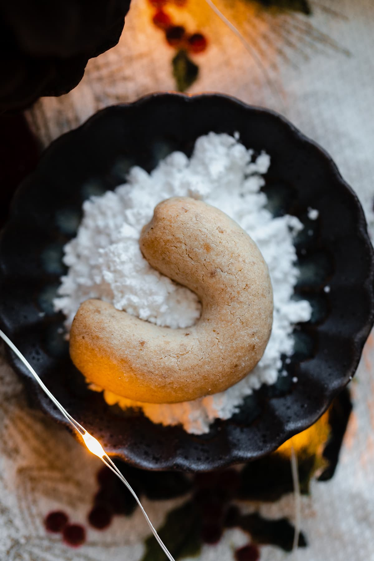 A single crescent cookie on a small plate with powdered white sugar and a string of fairy lights on the bottom left.