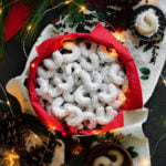 A square photo of Vanilla Walnut Crescent Cookies in a box with a red paper napkin surrounded with fairy lights, pine cones, and a pine branch