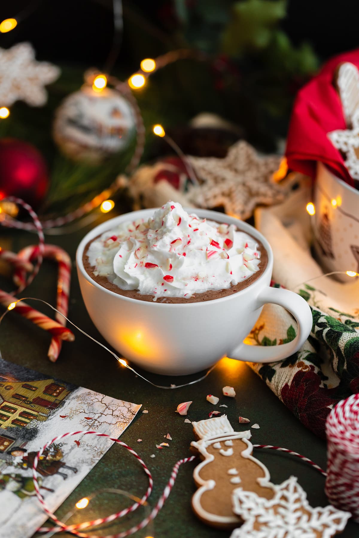 Peppermint hot chocolate in a white cup topped with whipped cream and crushed candy cane. Gingerbread cookies aranged around the cup. Dark green background.