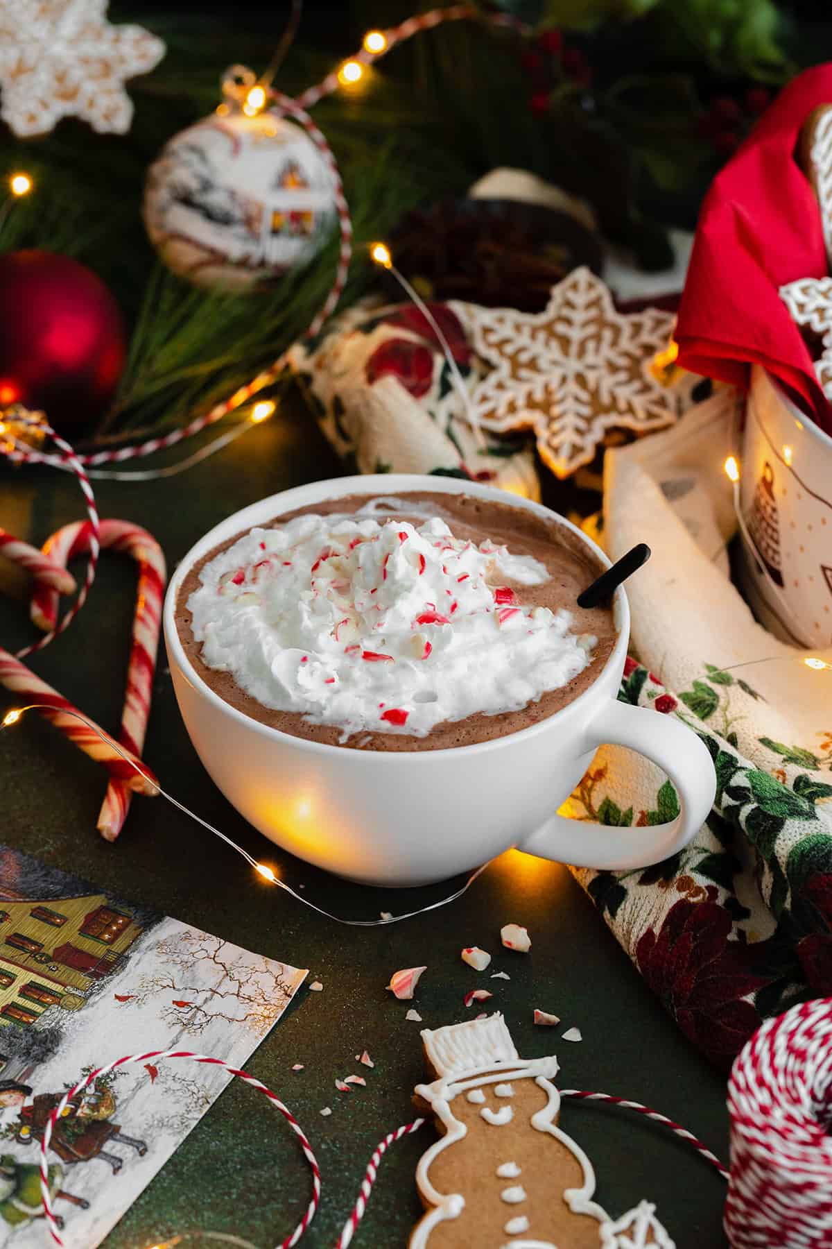 Peppermint hot chocolate in a white cup topped with whipped cream and crushed candy cane. Spoon inside the cup on the right side. Gingerbread cookies aranged around the cup. Dark green background.