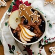 An overhead photo of Gingerbread oatmeal in a beige bowl garnished with sliced pear, a slice of apple, a few blueberries, and two gingrbread men. On light wooden background with a christmas tea towel under the bowl and gingerbread cookies around it.