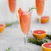 Grapefruit Mimosa in 3 flute glasses garnished with fresh rosemary and a quarter of a slice of grapefruit on the rim. Grapefruits as decoration in the background.