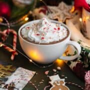 Peppermint hot chocolate in a white cup topped with whipped cream and crushed candy cane. Gingerbread cookies aranged around the cup. Dark green background.