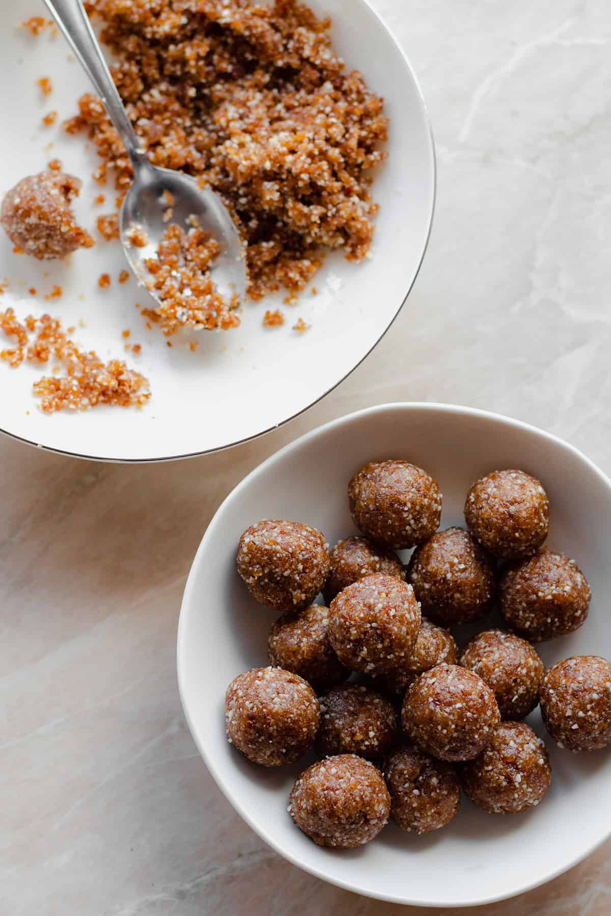 Energy balls in a white bowl on a marble counter. Another white bowl in the top left corner with the mixture before it's shaped into balls.