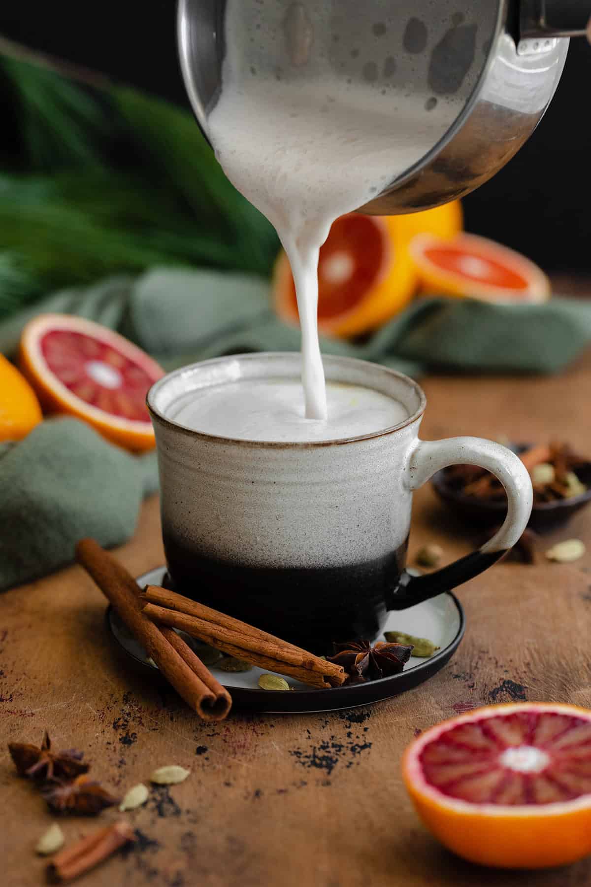 Frothed milk being poured into a mug on a light wooden table with blood oranges in the background.