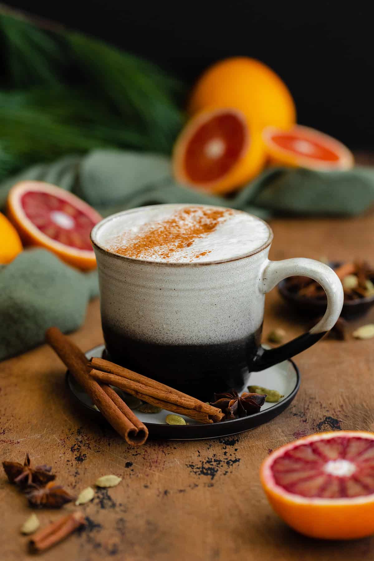 Blood Orange Chai Tea Latte in a ceramic black and white mug. On a light wooden table with blood oranges in the background.