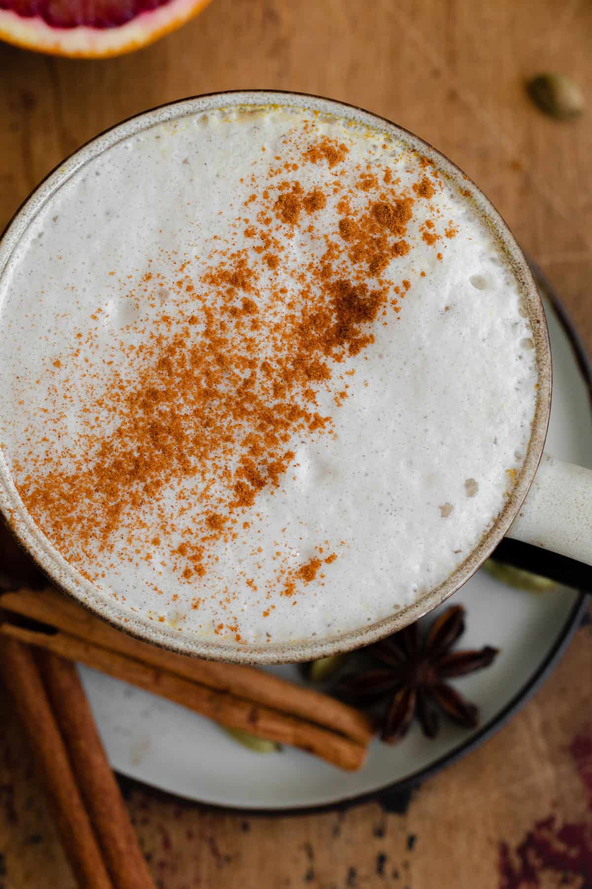 A close up of Blood Orange Chai Tea Latte in a ceramic mug on a small white plate. On a light wooden table.