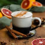 A square photo of Blood Orange Chai Tea Latte in a ceramic black and white mug. On a light wooden table with blood oranges in the background.