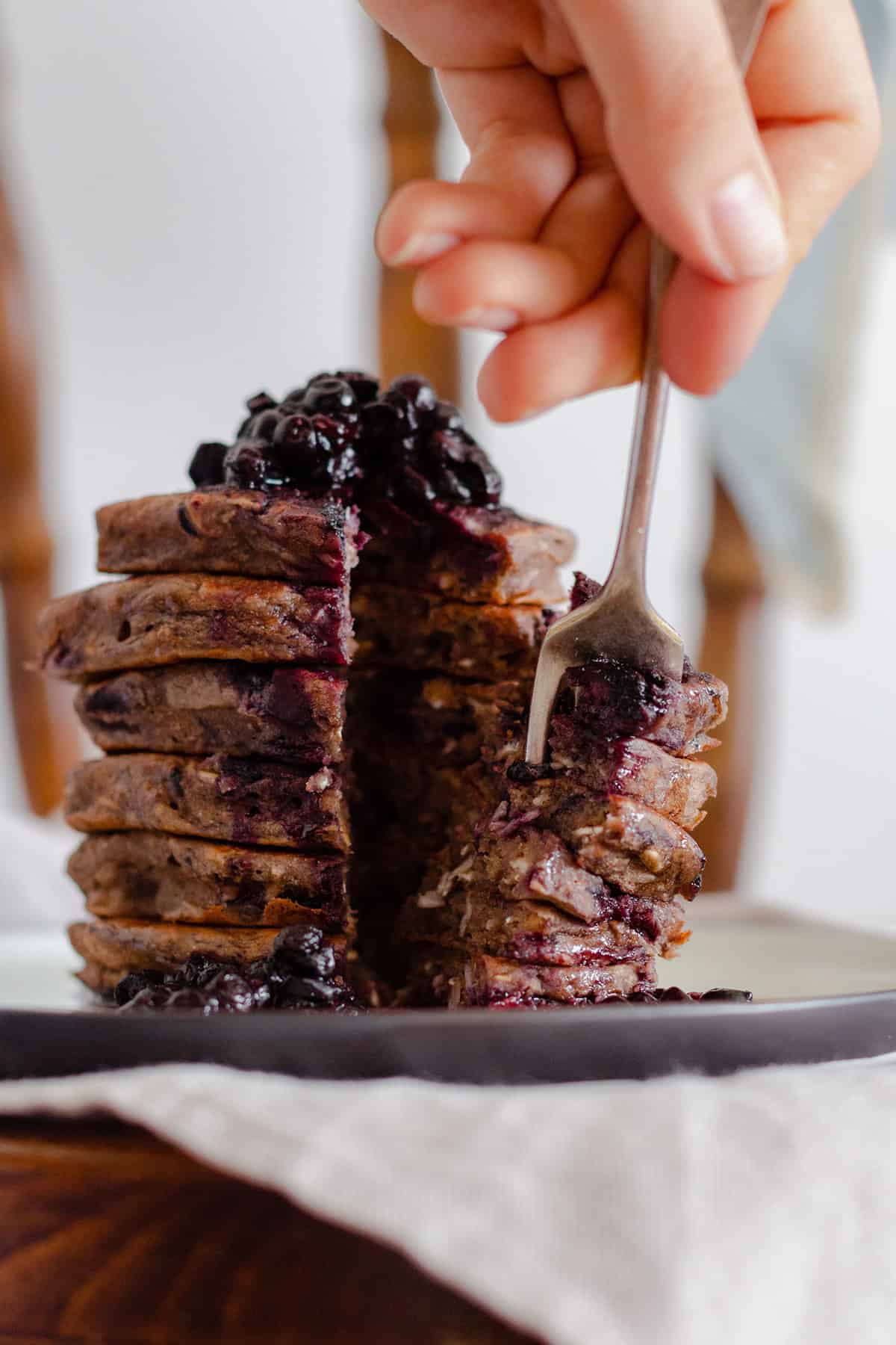 Buckwheat banana blueberry pancakes stacked on a beige plate on a chair. A hand scooping a piece of the layers with a fork.