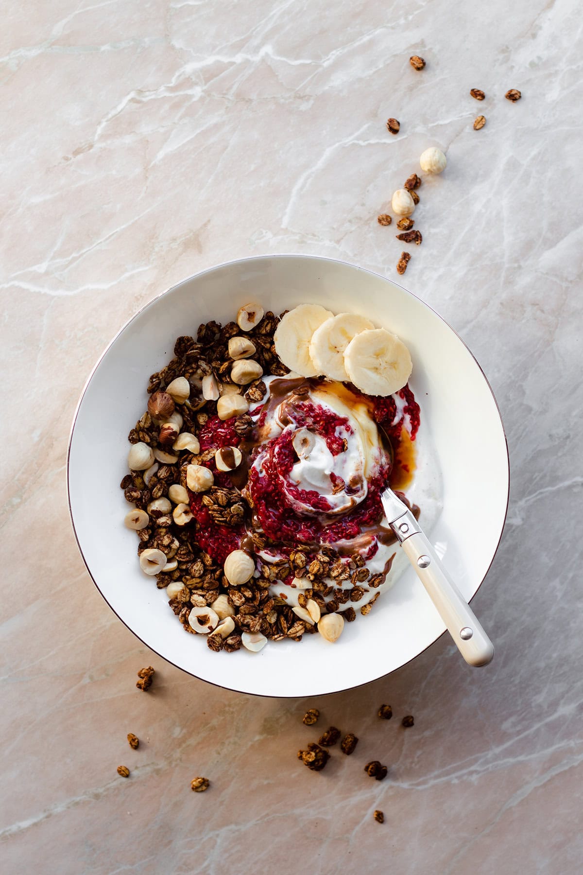 A white bowl on a marble countertop with carob granola, berry chia jam, maple syrup, and slices of banana.