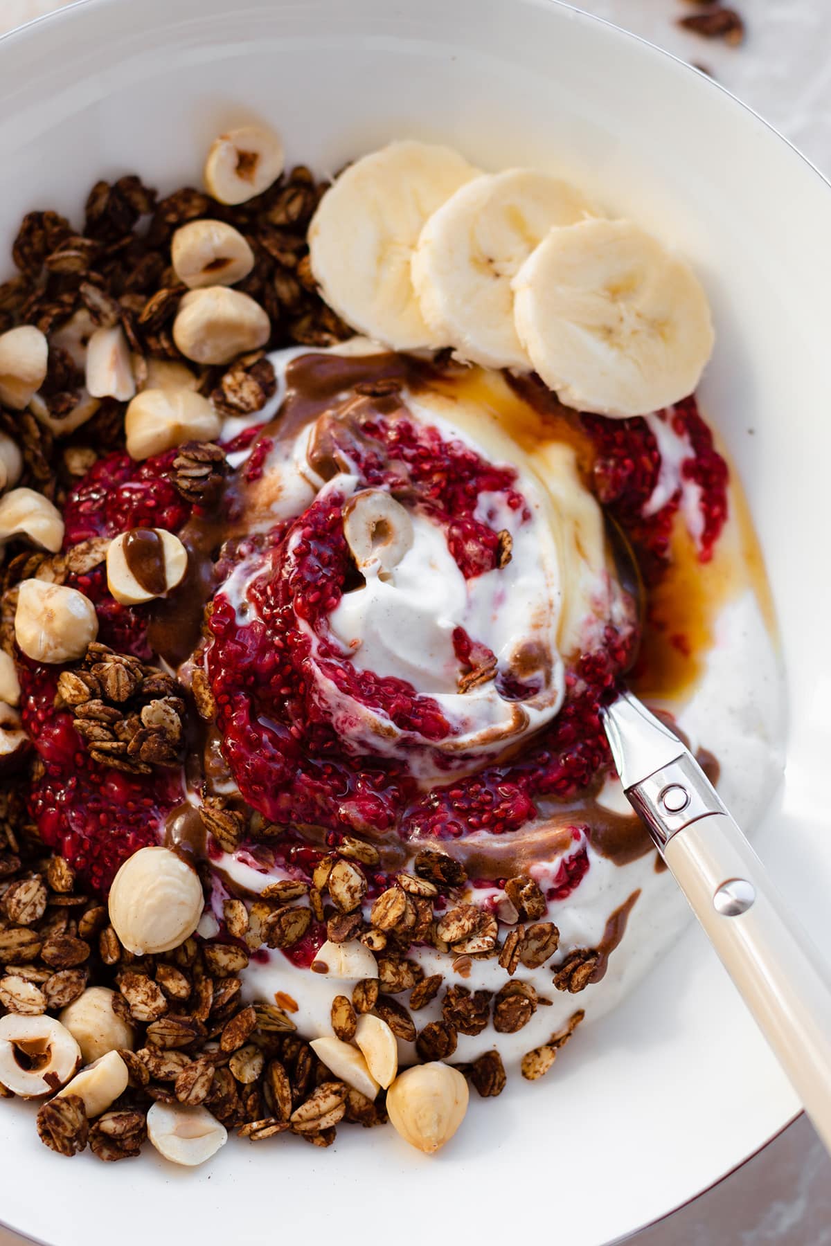 A close up shot of a white bowl on a marble countertop with carob granola, berry chia jam, maple syrup, and slices of banana.