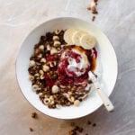 A white bowl on a marble countertop with carob granola, berry chia jam, maple syrup, and slices of banana.