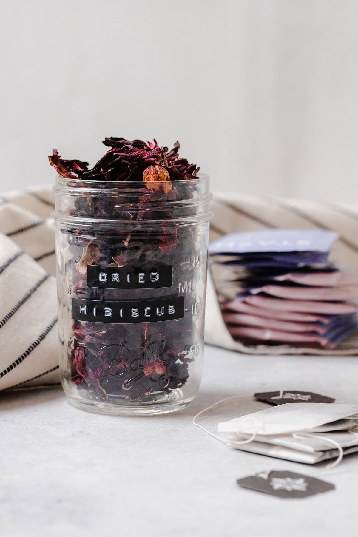 Dried hibiscus flower in a glass jar. On a light grey background with loose tea bags on the right side.