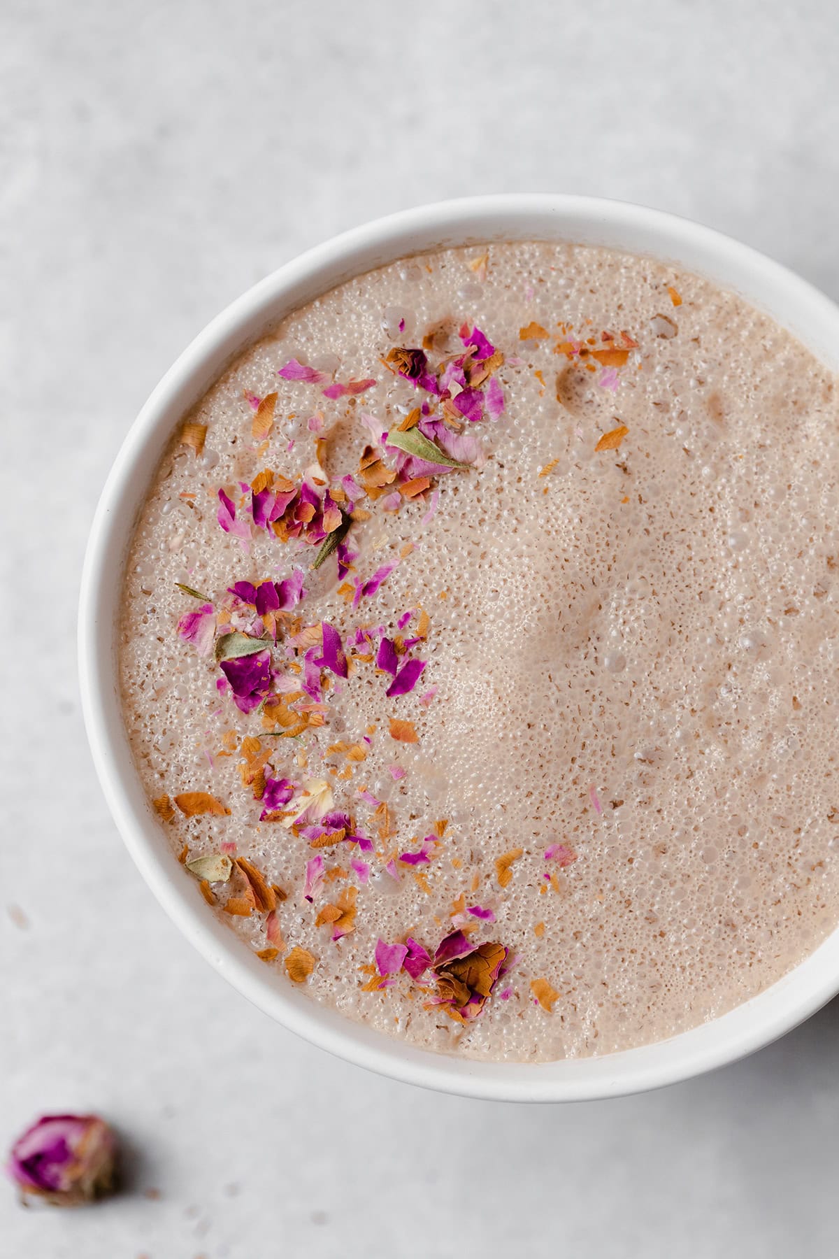 close up of hibiscus latte in a white mug on a light grey background. Garnished with crushed dried rose petals.