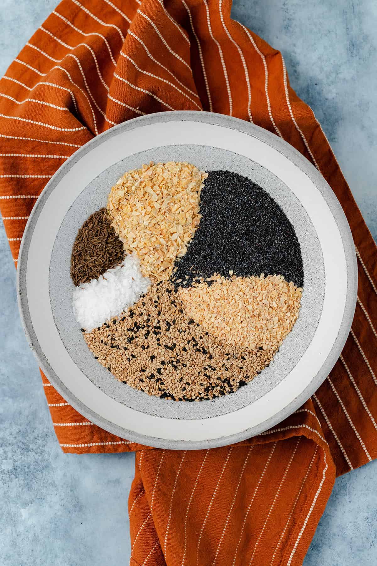 Everything bagel seasoning in low grey bowl on a blue table with an orange napkin under the bowl.