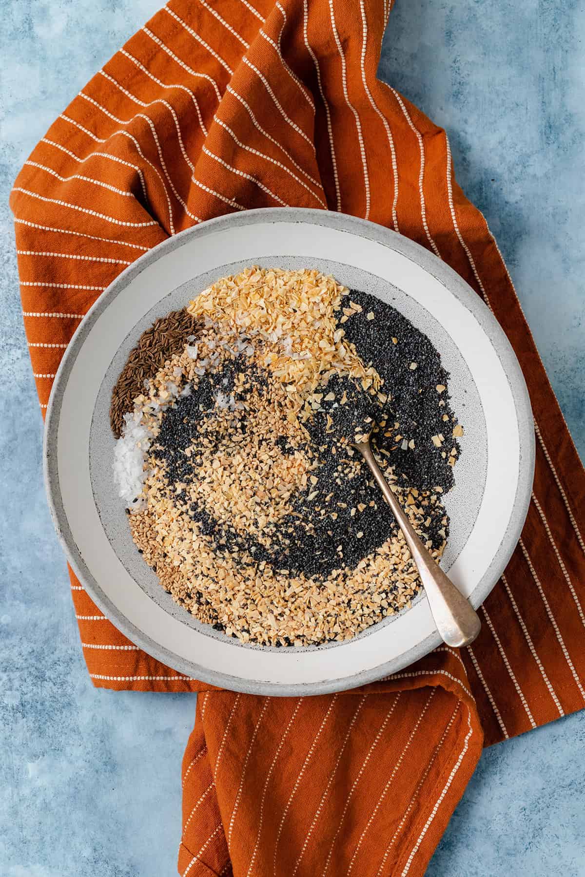 Everything bagel seasoning in low grey bowl on a blue table with an orange napkin under the bowl.