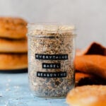 Everything bagel seasoning in a glass jar on a blue table with three bagels in the background and an orange napkin on the right.