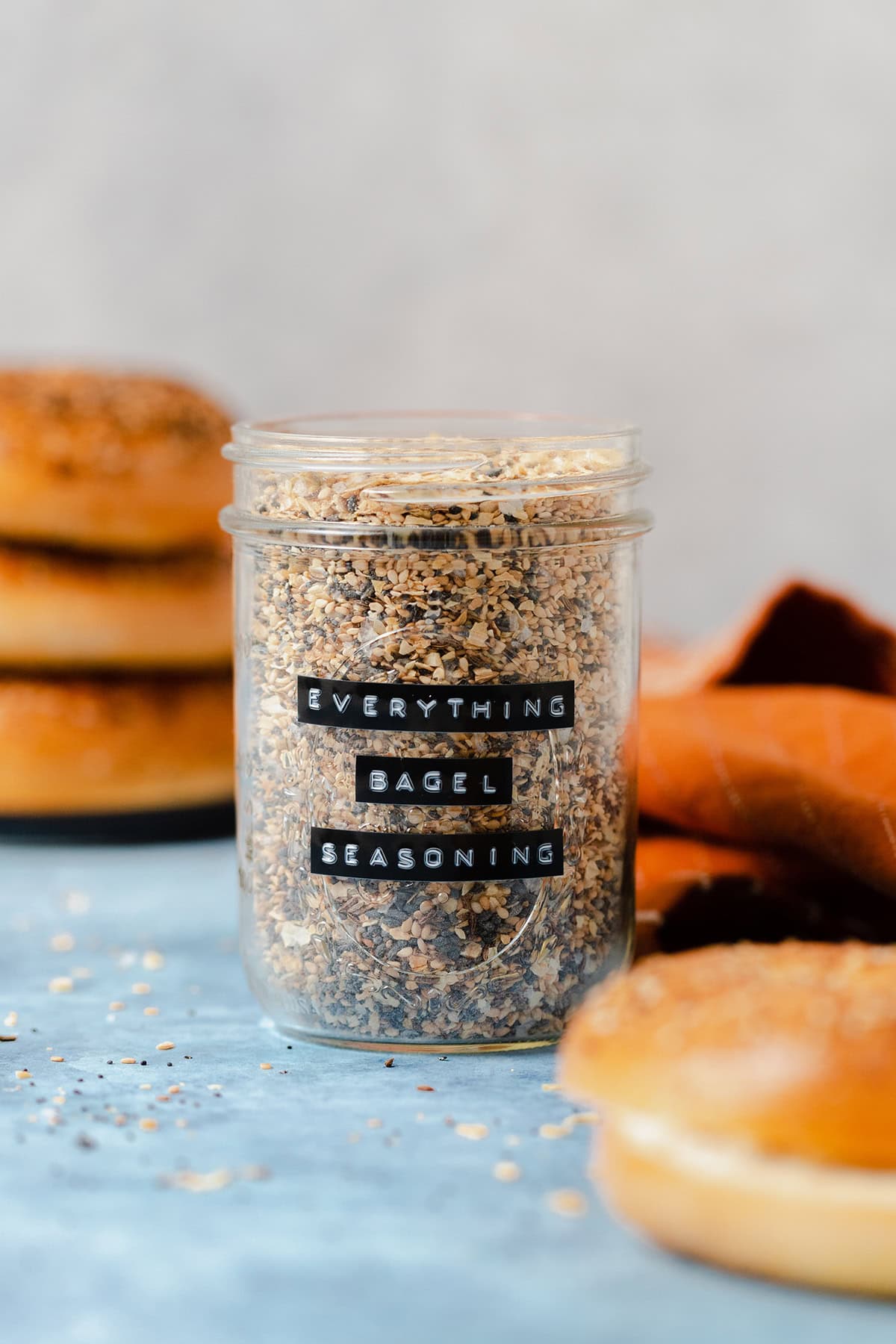 Everything bagel seasoning in a glass jar on a blue table with three bagels in the background and an orange napkin on the right.