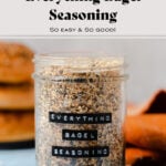 Everything bagel seasoning in a glass jar on a blue table with three bagels in the background and an orange napkin on the right.
