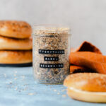 Everything bagel seasoning in a glass jar on a blue table with three bagels in the background and an orange napkin on the right.