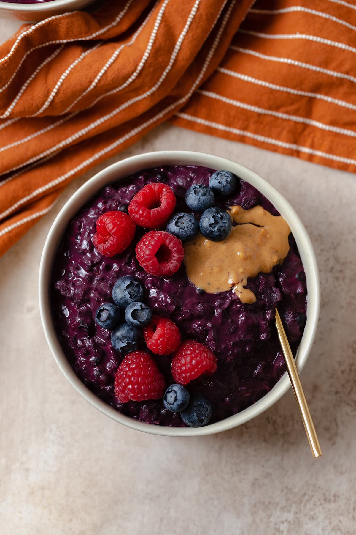 A beige bowl full of dark purple oatmeal topped with raspberries and blueberries. Gold spoon on the right side of the bowl.