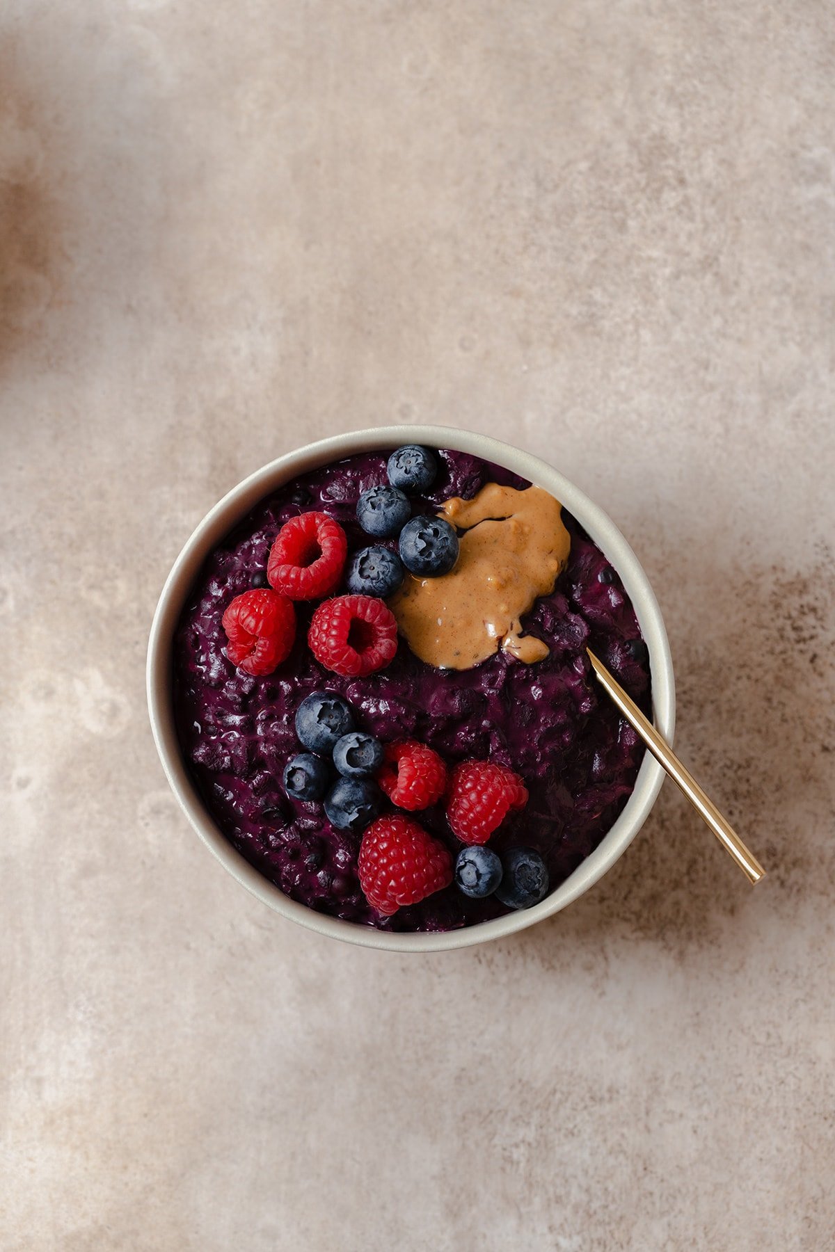 A photo of one beige bowl with a golden spoon on a beige background. Bowl full of purple maqui berry oatmeal.