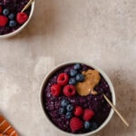 A photo of two beige bowls on a beige background. Bowls full of purple maqui berry oatmeal. Topped with raspberries, blueberries, and peanut butter.