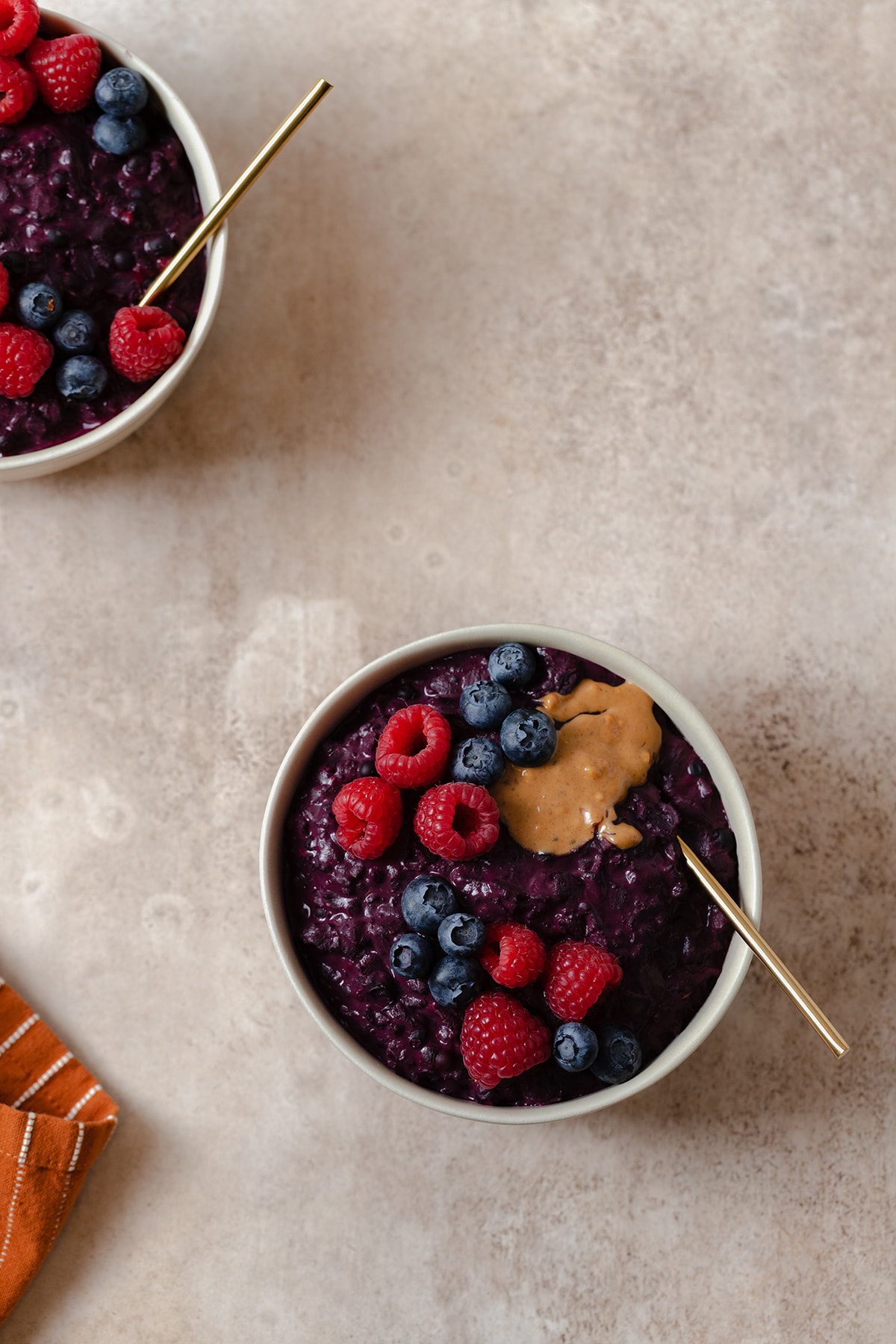 A photo of two beige bowls on a beige background. Bowls full of purple maqui berry oatmeal. Topped with raspberries, blueberries, and peanut butter.