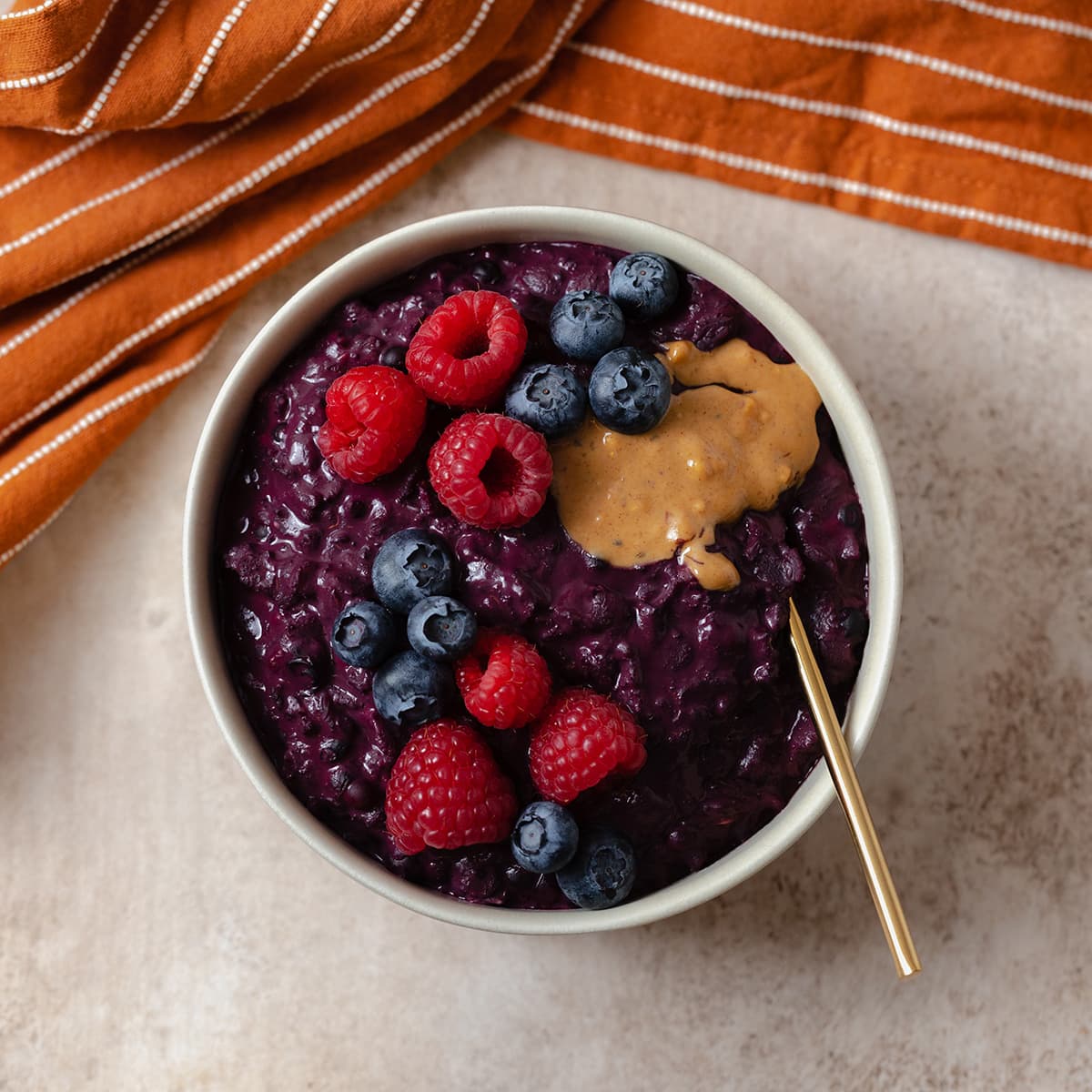 Deep purple oatmeal with maqui berry in a beige bowl on a light orange background. Served topped with raspberries, blueberries, and a spoonful of nut butter.