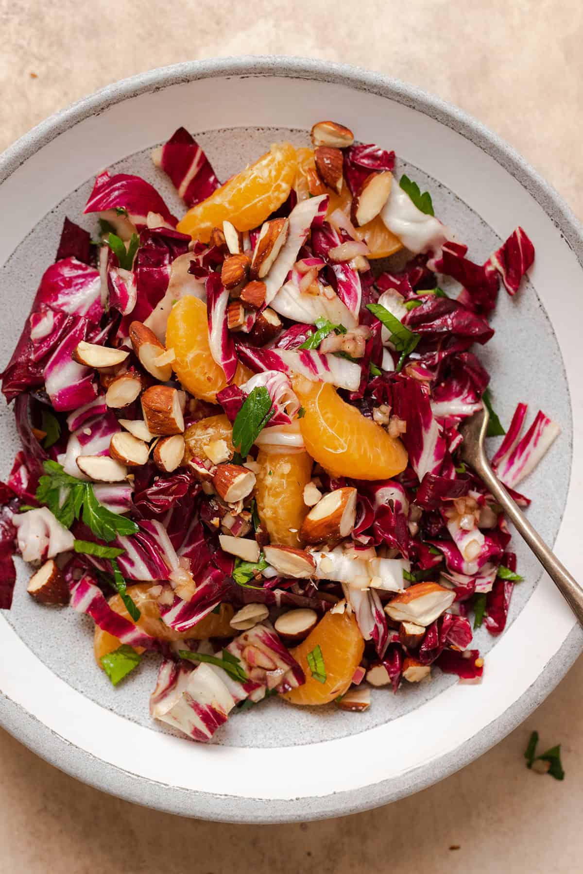 A close up shot of the Radicchio Clementine Salad shown on a shallow grey bowl with a white stripe along the edge. Fork resting on the right side of the bowl in the salad.