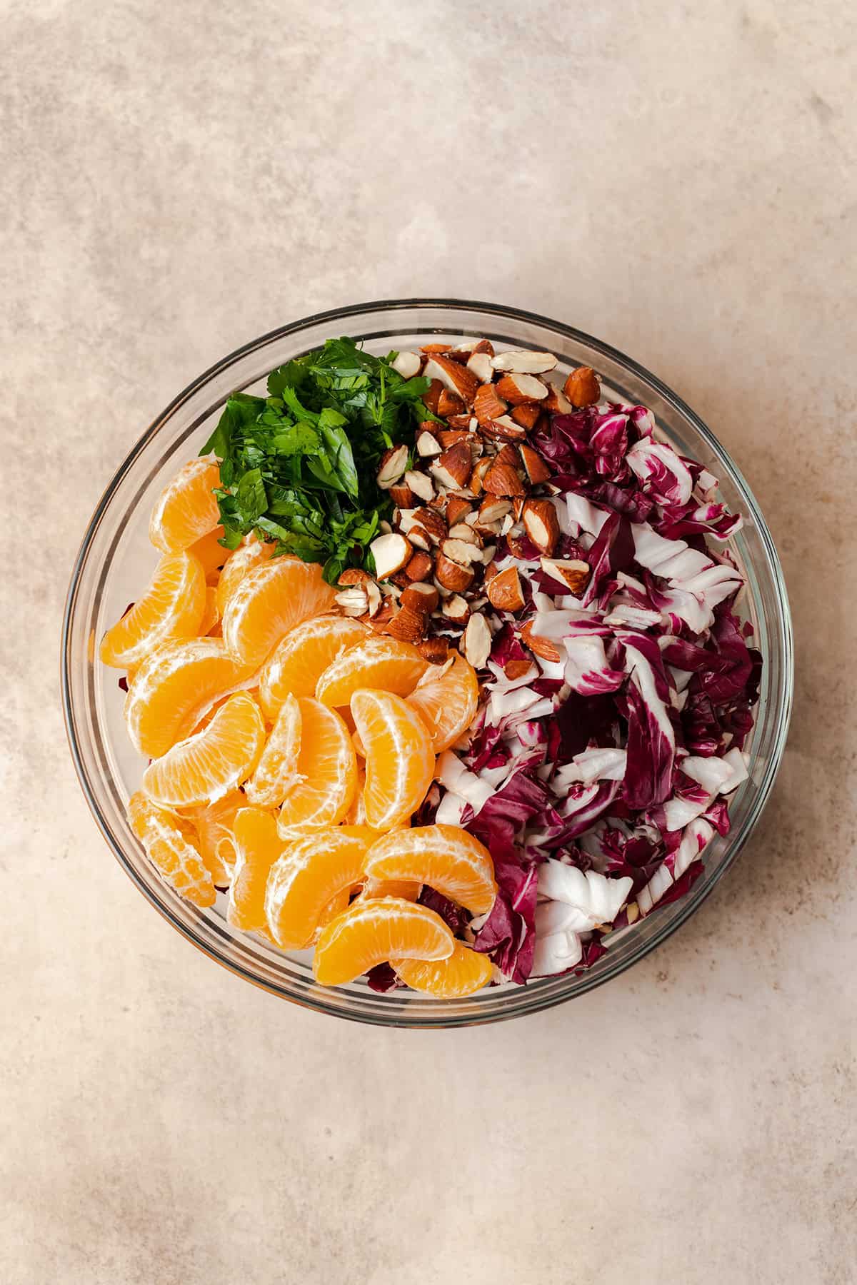 All ingredients for the clementine salad laid out in a glass bowl. There is chopped radicchio, chopped almonds, chopped parsley, and clementine pieces.