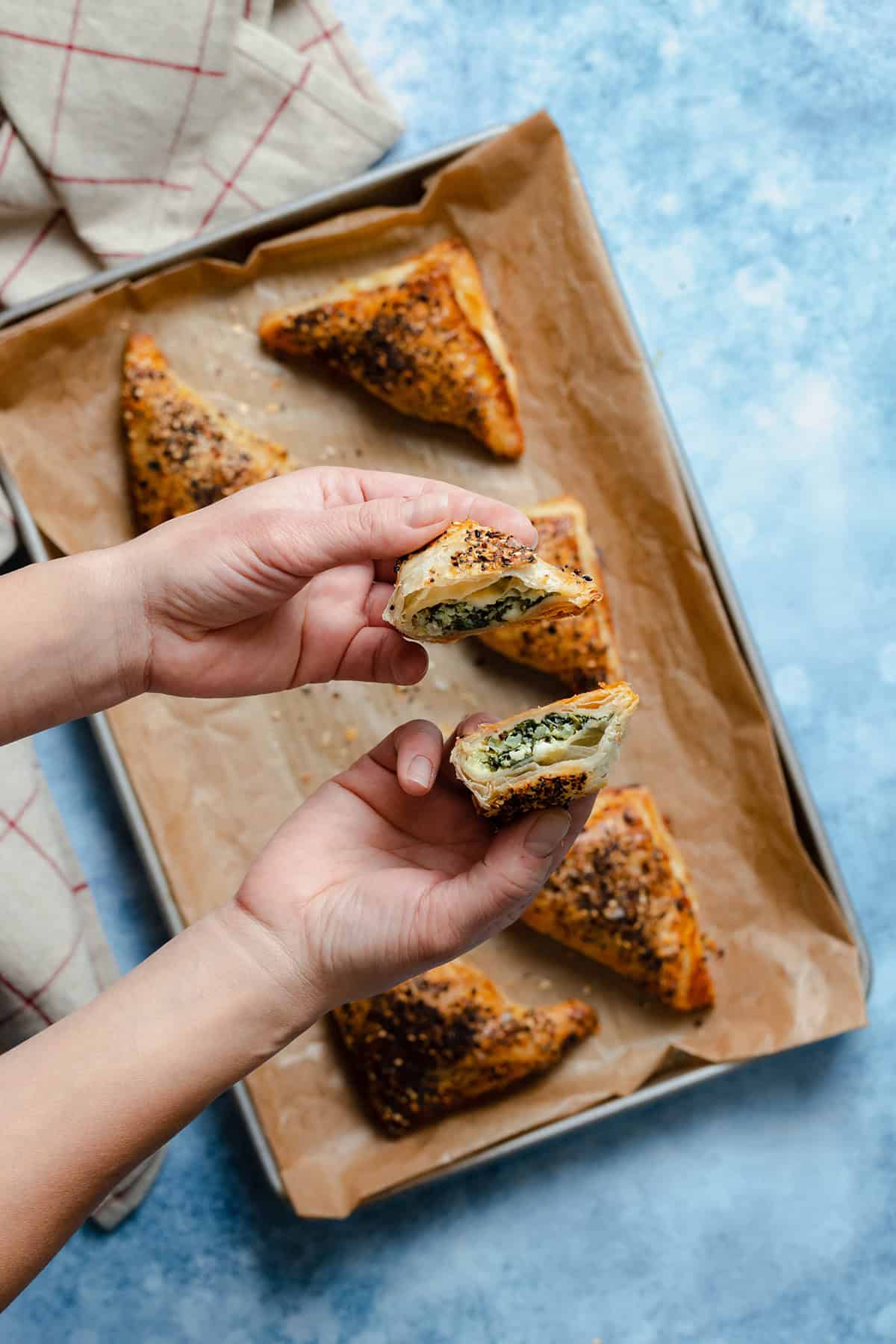 Spinach and Feta Puff Pastry Triangles on a baking sheet lined with parchment paper. Hands holding one triangle cut in half.