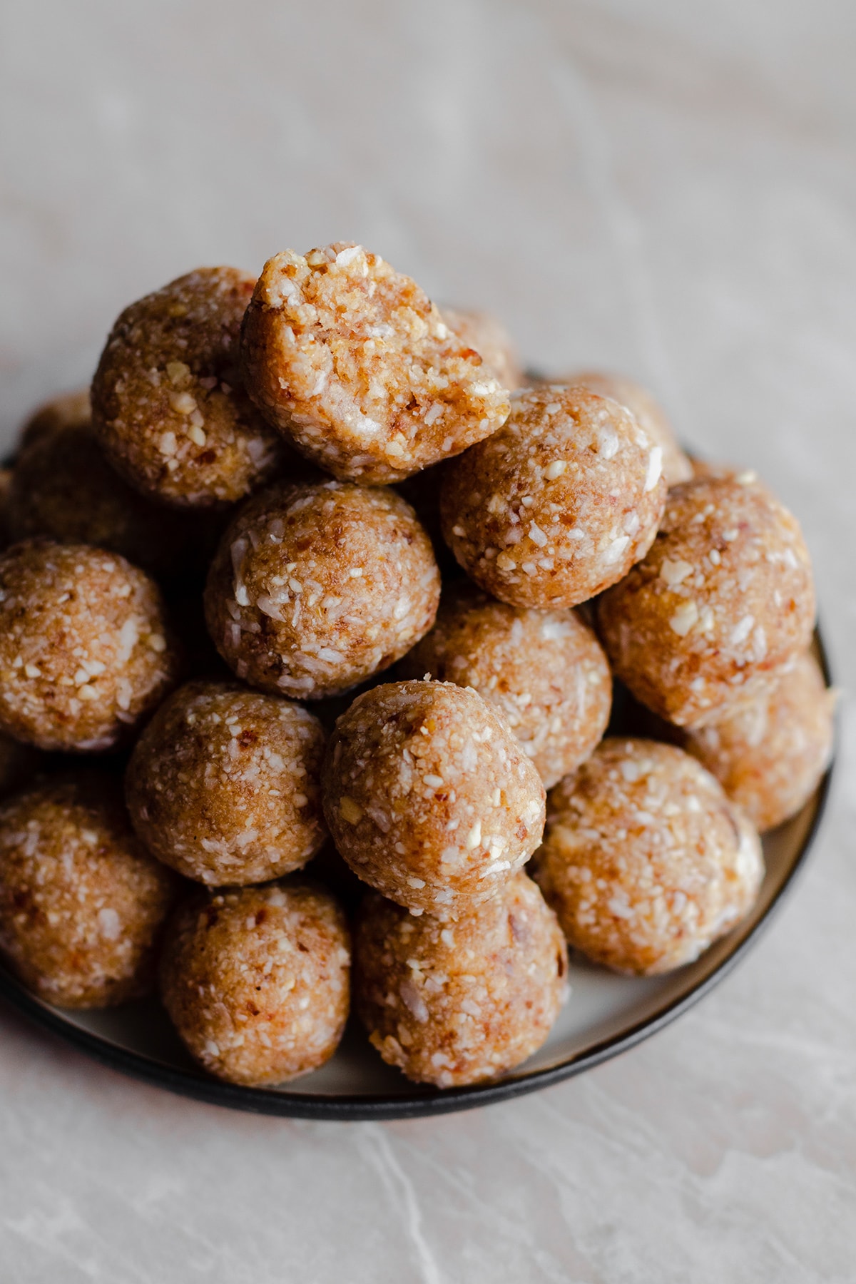 Coconut Energy Balls stacked up on a plate. Light marble background. With a bite taken out of the ball on the top.