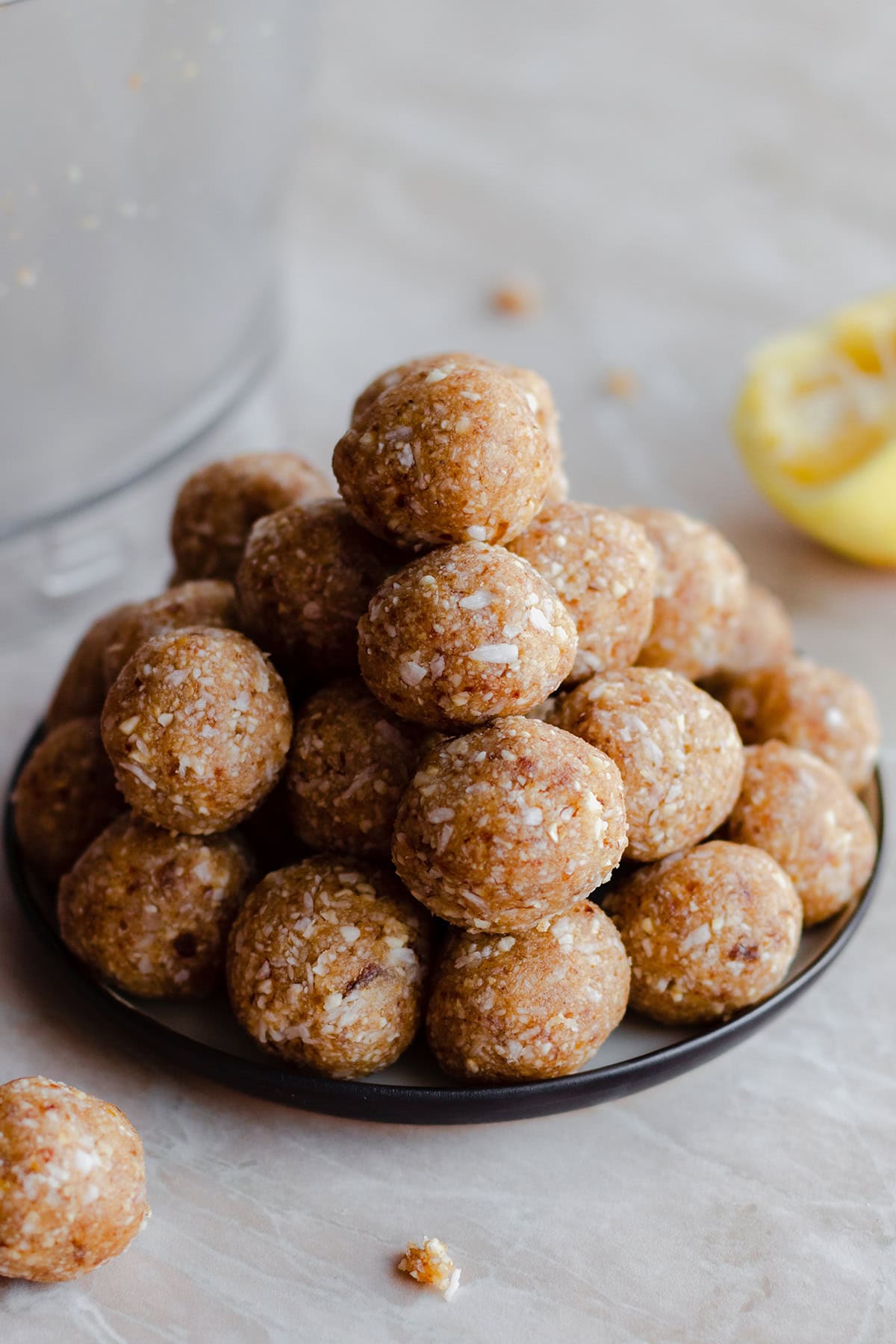 A photo of Zesty Lemon Coconut Energy Balls stacked up on a plate. Light marble background. Lemon in the background on the right side.