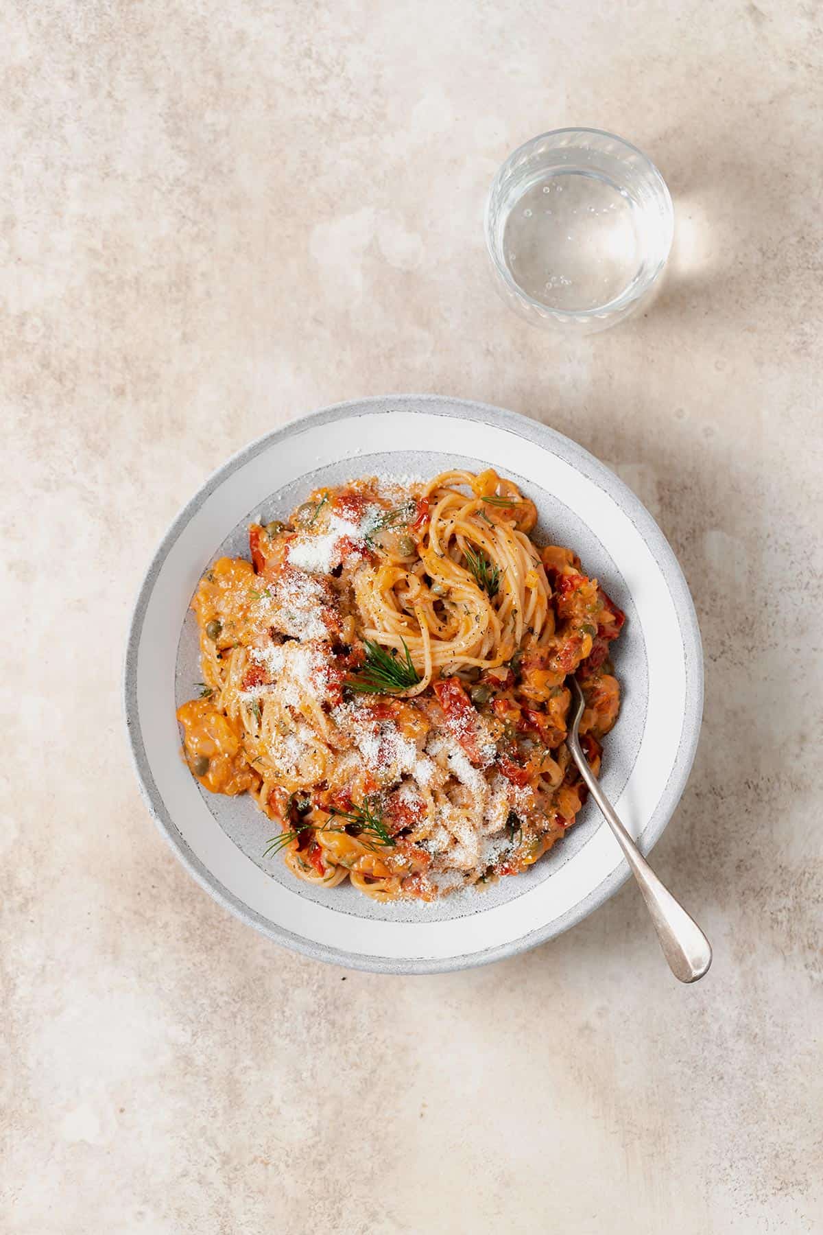 Creamy Sun-dried Tomato Pasta with Capers and Dill shown in a grey low bowl on a beige background. A glass with sparkling water on the right top side of the bowl.