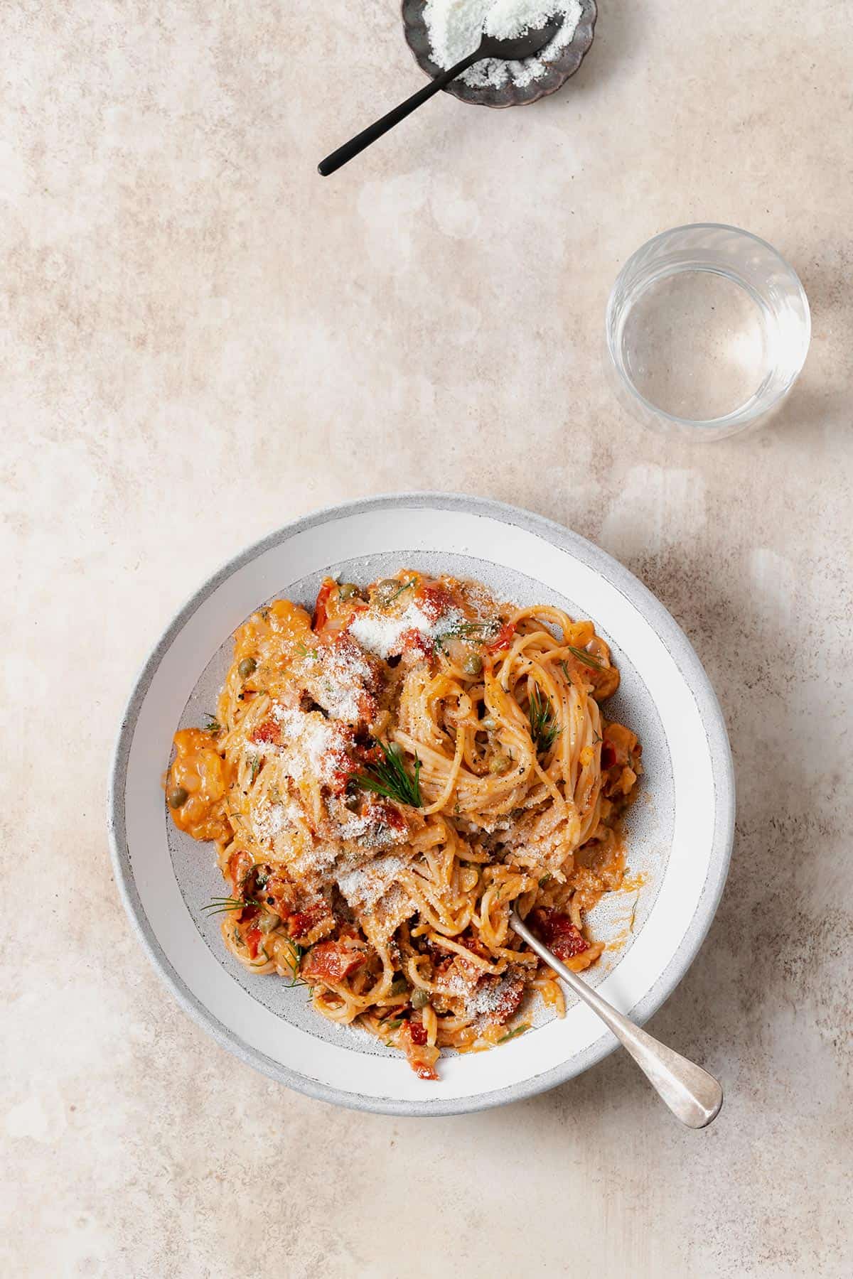 Creamy Sun-Dried Tomato Pasta with Capers, Dill, and a sprinkle of Pecorino shown in a grey low bowl on a beige background. A glass with sparkling water on the right top side of the bowl.