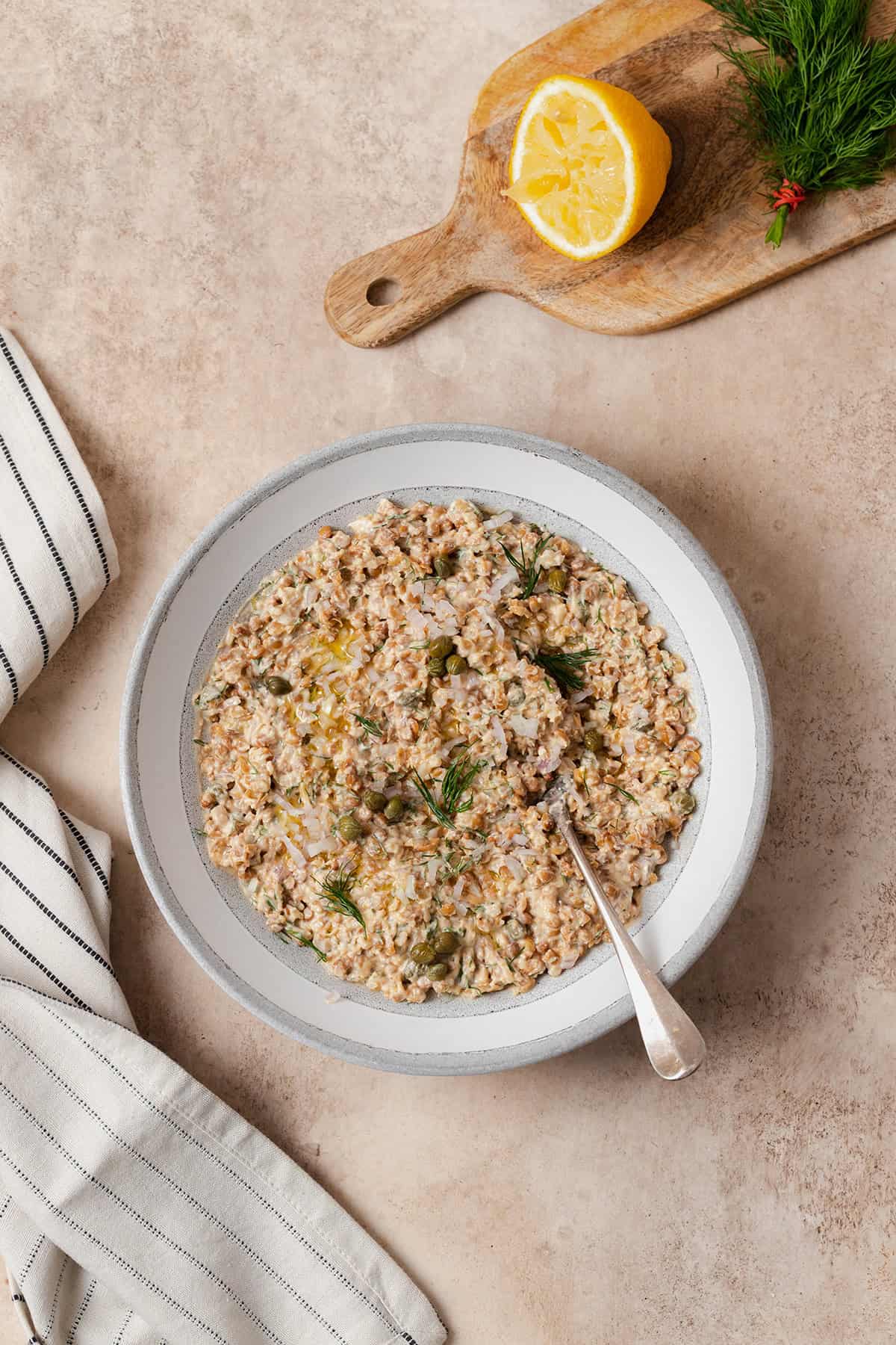 Lentil dip shown in a grey bowl with white strip along the edge. Spoon resting in the dip on the right side of the bowl. On a beige background with a black and white striped napkin on the left.