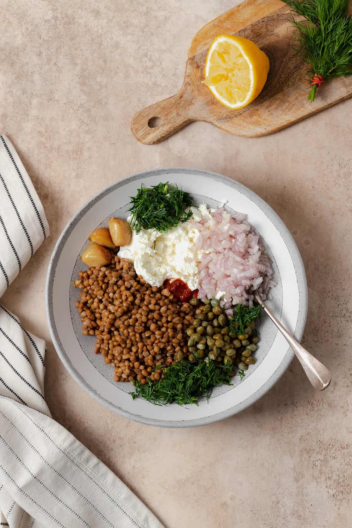 Ingredients shown in a grey bowl. Cooked brown lentils, capers, olive oil, half a lemon, a block of soft goat cheese, half a shallot, a bunch of fresh dill, roasted garlic, and garlic chili sauce.